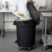 A chef in a professional kitchen putting food on a white tray into a black Rubbermaid BRUTE trash can.