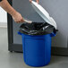A person putting a plastic bag into a blue Rubbermaid BRUTE recycling can with a white lid.