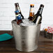A Tablecraft stainless steel beverage pail filled with ice and beer bottles on a table.