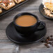 A black Libbey Driftstone porcelain cup filled with coffee on a saucer with pastries.