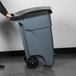 A person pushing a Rubbermaid grey wheeled trash can.