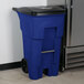 A blue Rubbermaid BRUTE rectangular trash can with wheels and a black lid next to a white refrigerator.