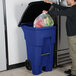 A man putting a plastic bag into a Rubbermaid blue wheeled rectangular trash can.