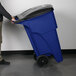 A man pushing a Rubbermaid blue wheeled rectangular trash can.