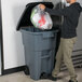 A man standing next to a Rubbermaid trash can with wheels holds a plastic bag and puts it in the trash can.