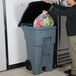 A man putting a plastic bag into a Rubbermaid 65 gallon gray wheeled rectangular trash can.