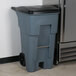 A Rubbermaid 65 gallon gray trash can with black lid next to a refrigerator.
