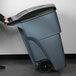 A person pushing a large gray Rubbermaid wheeled trash can.