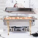A chef uses a Vollrath Classic Brass Trim electric chafing dish to cook food on a table at an outdoor catering event.