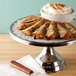 A Cal-Mil stainless steel cake stand with a cake and a cup of coffee on a table.