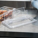 A clear plastic Cambro container lid with a spoon notch on top of a clear plastic container with food inside.
