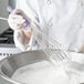 A person in a white coat using a Vollrath stainless steel piano whisk to stir a bowl of white liquid.