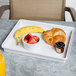 A white melamine box lid on a tray with a banana, strawberries, and a croissant.