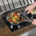 A person using a Vollrath Mirage Series drop in induction warmer to cook noodles and vegetables in a pan on a countertop.