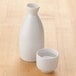 A white ceramic Town sake cup filled with liquid next to a white ceramic bottle.