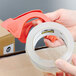 A hand holding a roll of 3M Scotch clear heavy-duty packaging tape with a tape dispenser.