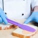 A person in blue gloves using a purple HS Inc. sandwich spreader to spread butter on a piece of bread.