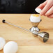 A hand using a Matfer Bourgeat stainless steel egg topper to remove the top of a soft boiled egg over a metal egg cup.