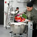 A man in a school kitchen using a Hobart vertical cutter mixer to chop vegetables in a large bowl.