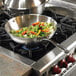 A Vollrath Miramar stainless steel stir fry pan with vegetables on a stove.