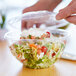A person using a clear plastic Eco-Products salad bowl with a lid to hold a salad.