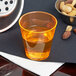 A Fineline Quenchers neon orange plastic shot cup filled with orange liquid on a table with a bowl of nuts.