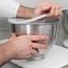 A person's hands using a white plastic KitchenAid mixer bowl cover to mix ingredients in a bowl.