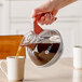 A person pouring coffee into a Bunn coffee decanter with an orange handle.
