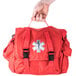 A hand holding a red Medi-First emergency kit with a black cross on it.