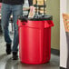 A man standing next to a Rubbermaid red trash can and putting a bag in it.