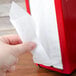 A hand pulling a white napkin from a red Tablecraft tallfold napkin dispenser.