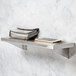 A Bobrick stainless steel wall mount shelf with a purse and phone on it.