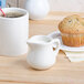A white Tuxton China creamer on a table with a cup of coffee and a muffin.