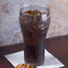 A clear plastic pebbled soda glass filled with dark soda and ice on a table with a napkin and food.