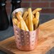 A cup of french fries in a hammered copper french fry cup on a wooden cutting board.