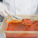 A gloved hand uses a Vollrath ivory Spoodle to scoop red sauce into a glass container.