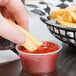 A hand holding a french fry and dipping it into a clear plastic container of ketchup.