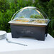 A black Sterno chafing dish with clear dome lid covering food on a table with plates.