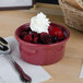 A red Tuxton casserole dish filled with a bowl of fruit and whipped cream with a spoon.