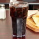 A Carlisle clear plastic tumbler filled with soda and ice on a table with chips and salsa.