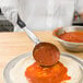 A person using a Vollrath black round portion spoon to put red sauce on pizza dough.