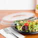 A salad in a Genpak clear dome lid on a table with a fork and knife.