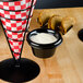 A black Tablecraft melamine ramekin with white liquid in it on a table with a red and white checkered wrapper in a black metal holder.