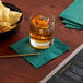 A glass of liquid with ice and a bowl of potato chips on a table with a Choice hunter green cocktail napkin.