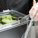 A person in gloves uses a Carlisle Clear plastic lid with a notch to cover a food container on a salad bar counter.