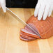 A person using a Mercer Culinary Renaissance forged carving knife to slice ham on a cutting board.