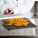 A person in a white coat cooking french fries using a Baker's Mark loose weave mesh screen on a black counter.