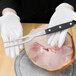 A person in white gloves holding a Mercer Culinary Renaissance forged carving fork over a ham.