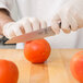 A person in white gloves using a Mercer Culinary Z&#252;M&#174; forged utility knife to slice a tomato on a wooden table.