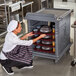 A woman in a chef's uniform kneeling in front of a Granite Gray Cambro meal delivery cart and putting dishes inside.